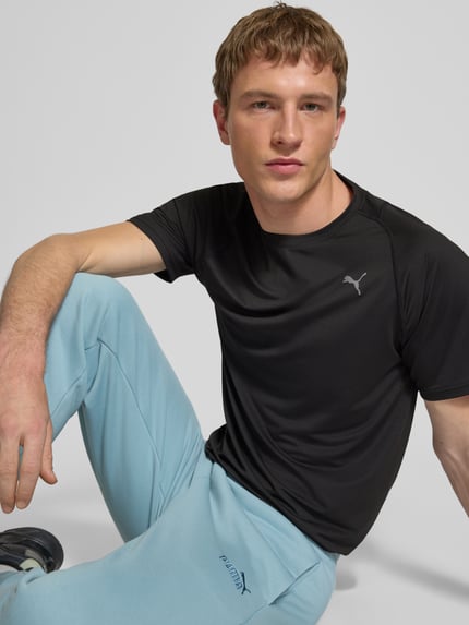 Męski model nosi T-shirt mit Label-Print von PUMA PERFORMANCE, mieszanka materiałów z dodatkiem streczu, krój regular fit, prążkowany okrągły dekolt, długość tyłu w rozmiarze M: 71 cm, kolor czarny, faktura gładka, z przodu nadruk z logo, ujęcie: zbliżenie na górną część ciała