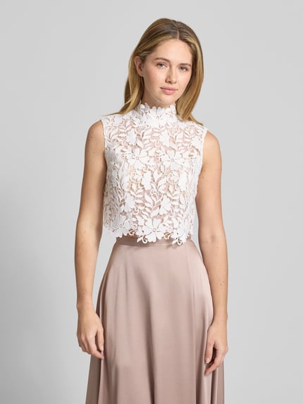 Frauenmodel trägt Regular Fit Top in semitransparentem Design von Luxuar mit Allover-Spitzen-Optik, Stehkragen, Ärmelloses-Design, integrierte Pads, Farbe weiß, Vorderansicht, Nahaufnahme Oberkörper.