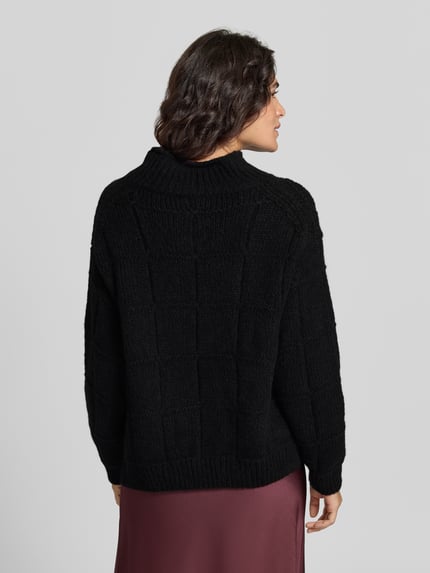 Frauenmodel trägt czarny sweter wełniany z dzianiny o pudełkowym kroju z wysokim kołnierzem typu mock neck, długi rękaw, jednokolorowy, imitacja dzianiny, szwy działowe, prążkowane zakończenia rękawów i dołu, sweter aus Wolle mit Strukturstrick von Marc Cain, Ganzkörperansicht Rückansicht.