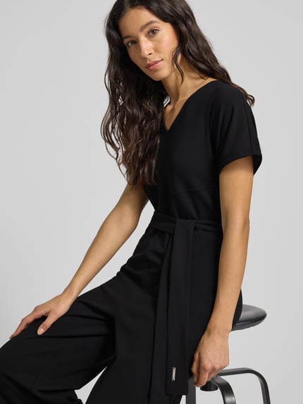 Frauenmodel trägt Jumpsuit mit V-Ausschnitt von Calvin Klein Womenswear, schwarzer Material-Mix mit Stretch-Anteil, Taillengürtel, Nahaufnahme Oberkörper.