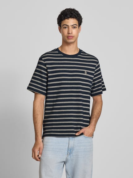 Męski model nosi Strukturiertes T-Shirt mit Logostickerei auf der Brust od REVIEW z fakturowanym wzorem, 100% bawełna; prosty krój, okrągły dekolt, wyhaftowane logo i haft angielski na piersi, długość tyłu w rozmiarze m: 70 cm, kolor granatowo-beżowy; widok całej sylwetki z przodu.