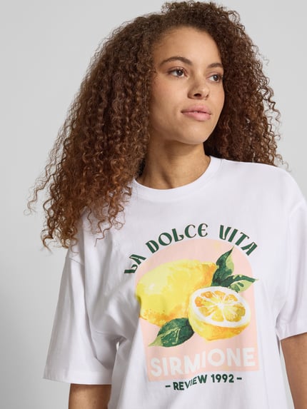 Modelka kobieca ma na sobie damski t-shirt z nadrukiem z motywem na plecach 'T-Shirt mit rückseitigem Motiv-Print' od REVIEW, wykonany z czystej bawełny, krój oversized, okrągły dekolt, długość tyłu w rozmiarze S: 68 cm, zbliżenie górnej części ciała z przodu.