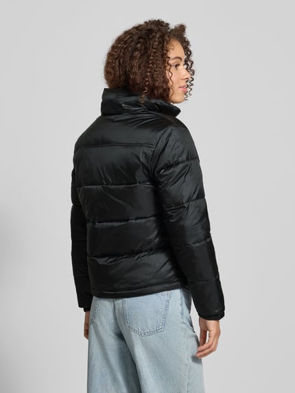 Frauenmodel trägt Regular Fit Steppjacke mit verdeckter Kapuze von Tommy Jeans in Schwarz, glatte Stepp-Textur, Ganzkörperansicht Rückansicht.