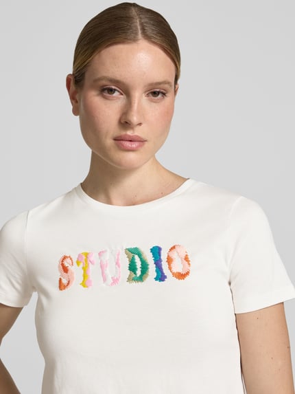 Modelka nosi Regular Fit T-Shirt mit Logo-Stitching od JAKE*S STUDIO WOMAN, biała bawełna, okrągły dekolt, przód haft kolorowy, zbliżenie na górną część ciała.