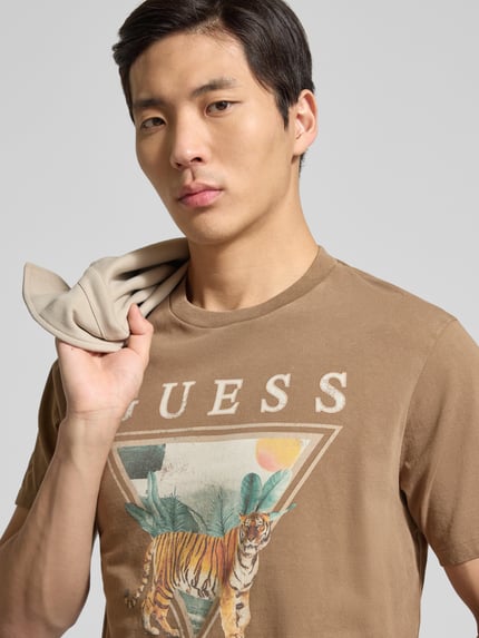 Mężczyzna model nosi T-Shirt z motywem nadruku model 'TIGER SPIRIT' od Guess, brązowe kolory, bawełna, krój regular fit, zbliżenie na górną część ciała.