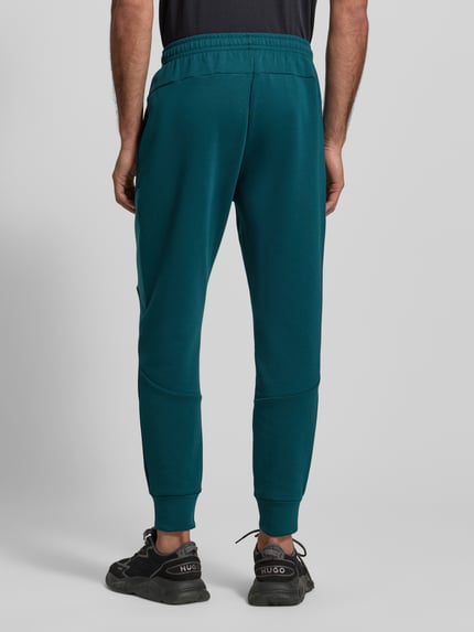 Mężczyzna nosi Spodnie dresowe Regular Fit Sweatpants mit Logo-Print Modell 'Unstoppable' von Under Armour, elastyczna mieszanka materiałów z dodatkiem bawełny, krój regular fit, kieszeń na nogawce z zamkiem błyskawicznym, elastyczny pas i zakończenia nogawek, długość wewnętrzna nogawki w rozmiarze M: 73 cm, widok całej sylwetki z tyłu.