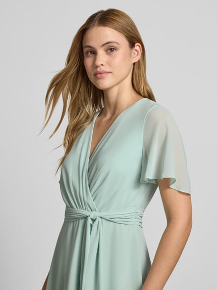 Frauenmodel trägt Knielanges Cocktailkleid in Wickel-Optik von Christian Berg Cocktail aus mintfarbenem Chiffon mit V-Ausschnitt, Bindegürtel, A-Linie, Nahaufnahme Oberkörper. Rückenlänge bei Größe 36: 100 cm, Brustweite: 33 cm, Ärmellänge: 22 cm.