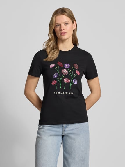 Frauenmodel trägt Slim Fit T-Shirt aus reiner Baumwolle Modell 'KITA' von Only, schwarzer Jersey, Blumen-Print, Pailletten, Ganzkörperansicht von vorne.