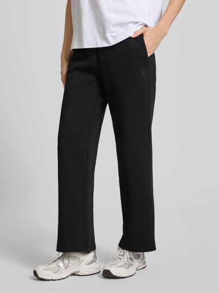 Kobiece modelka nosi Sweatpants mit elastischem Bund und Logo-Print od ARMANI EXCHANGE, krój straight fit, elastyczny pas, nadruk z logo, wpuszczane kieszenie, długość wewnętrzna nogawki w rozmiarze S: 72 cm, czarny kolor, gładka tekstura, widoczny biały t-shirt oraz sportowe buty, całkowity widok z przodu.