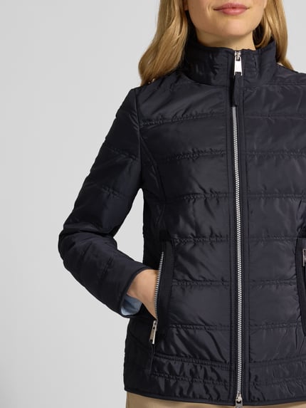Modelka nosi czarną Steppjacke mit Stehkragen und Reißverschlusstaschen od Fuchs Schmitt, krój slim fit, dwukierunkowy zamek, boczne kieszenie na zamek, detal z logo, długość tyłu w rozmiarze 36: 65 cm, szerokość ramion w rozmiarze 36: 45 cm, szerokość w biuście w rozmiarze 36: 42 cm, długość rękawa w rozmiarze 36: 65 cm, stonowany połysk, pikowana faktura, zbliżenie na górną część ciała.