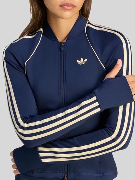 Frauenmodel trägt dunkelblaue Slim Fit Trainingsjacke mit Zweiwege-Reißverschluss Modell 'OG SP MIDLAY' von adidas Training, cremefarbene Streifen, Nahaufnahme Oberkörper.