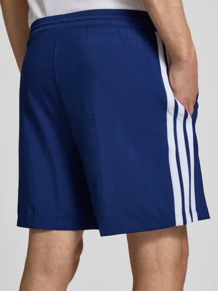 Männermodel trägt Regular Fit Sport-Shorts mit elastischem Bund von adidas Sportswear, unifarben blau mit weißen Kontraststreifen, Ganzkörperansicht Rückansicht.