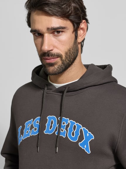 Männermodel trägt Hoodie mit Label-Print Modell 'Brady' von Les Deux in Dunkelbraun aus reiner Baumwolle mit Känguru-Tasche, Nahaufnahme Oberkörper. Regular-Fit, Rückenlänge 67 cm, Ärmellänge 72 cm, blauer Schriftzug.