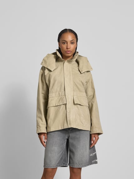 Kobiece model nosi kurtka 'Jacke aus reiner Baumwolle mit Kapuze' od Victoria Beckham w kolorze beżowym, luźny krój, bawełna, kaptur, wpuszczane kieszenie, zamek błyskawiczny i listwa guzikowa, długość tyłu w rozmiarze XS/S: 67 cm, szerokość w rozmiarze XS/S: 74 cm, długość rękawów w rozmiarze XS/S: 56 cm, widok całej sylwetki z przodu.