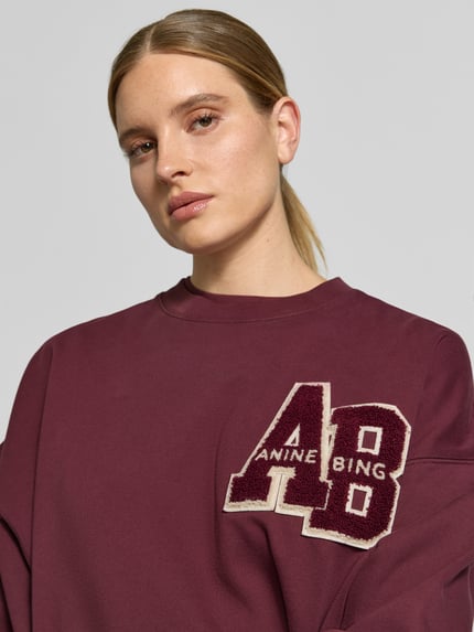 Kobieta modelka nosi Oversized Sweatshirt aus Baumwolle od Anine Bing, bordowa bluza z bawełny z detalem z logo, okrągły dekolt, elastyczne wykończenia, długość tyłu w rozmiarze S: 61 cm, zbliżenie na górną część ciała