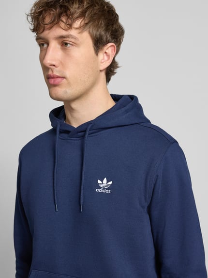 Männermodel trägt Hoodie mit Kapuze von adidas Originals in blauw, Katoenmix, los model, capuchon met tunnelkoord, labelstitching, kangoeroezak, ribboorden, ruglengte maat M 68 cm, mouwlengte maat M 69 cm, Nahaufnahme Oberkörper.