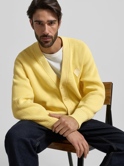 Männermodel draagt Relaxed Fit Gebreid jack uit katoenmix model 'COTES PERLEES' van Lacoste in effen geel met knoopsluiting, labelopschrift en labelstitching, V-hals, lange mouw, geribde mouwboord en rompzoom, materiaalverwerking: knitwear, katoenmix. Nahaufnahme Oberkörper.