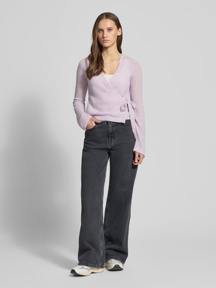 Frauenmodel trägt Strick-Cardigan in Wickeloptik von Review in zartem Flieder, kombiniert mit weiter Jeans, Ganzkörperansicht von vorne. Rückenlänge 50 cm, Ärmellänge 66 cm, Schleifenverschluss, V-Ausschnitt und langer Ärmel, Regular Fit.
