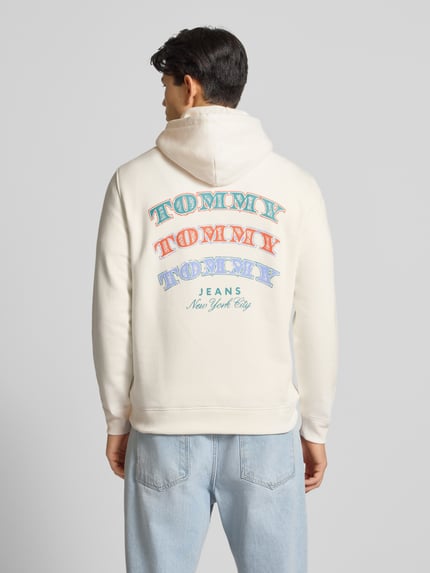 Mężczyzna nosi Regular Fit Hoodie aus reiner Baumwolle von Tommy Jeans, kremowa bluza z kolorowym logo, długi rękaw, raglan, kieszeń kangurka, bawełna, widok z tyłu całej sylwetki.