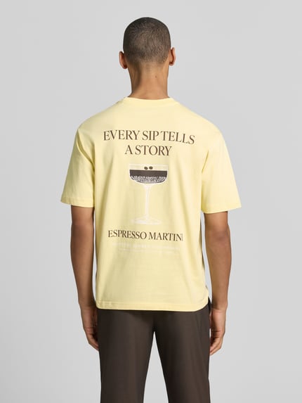 Männermodel trägt T-Shirt mit rückseitigem Motiv-Print von MCNEAL in Gelb, Rundhalsausschnitt, Statement-Print, Rückenlänge 70 cm, Ganzkörperansicht Rückansicht.