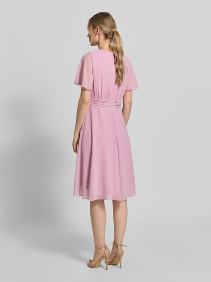 Frauenmodel trägt Knielanges Cocktailkleid in Wickel-Optik von Christian Berg Cocktail aus rosa Chiffon mit Bindegürtel, A-Linie, Ganzkörperansicht Rückansicht