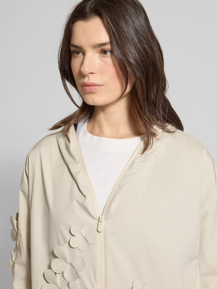 Frauenmodel trägt Regular Fit Regenjacke mit floralen Applikationen Modell 'Rosa' von SAVE THE DUCK in Beige, Logo-Patch, Reißverschluss, Nahaufnahme Oberkörper. Rückenlänge 72 cm, Schulterbreite 57 cm, Brustweite 45 cm, Ärmellänge 50 cm.