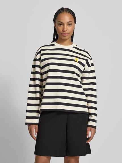 Kobiece model nosi Oversized Sweatshirt aus reiner Baumwolle Modell 'FRANKAA BOLD' od Armedangels, dwukolorowy wzór w paski, aplikacja z metką, wyhaftowane logo, okrągły dekolt, długi rękaw, dzianina dresowa z czystej bawełny, widok całej sylwetki z przodu.