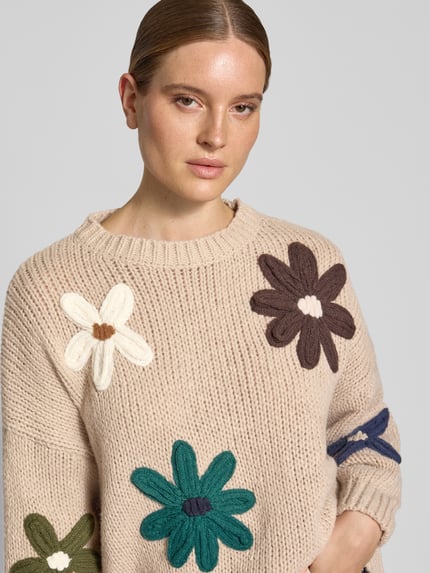 Kobieta modelka nosi Relaxed Fit sweter z dzianiny z kwiatowym haftem od Apricot, imitacja dzianiny, długi rękaw, okrągły dekolt, prążkowane wykończenia, beżowy kolor, kolorowe aplikacje kwiatowe, rozmiar S: długość tyłu 57 cm, długość rękawa 44 cm, zbliżenie na górną część ciała