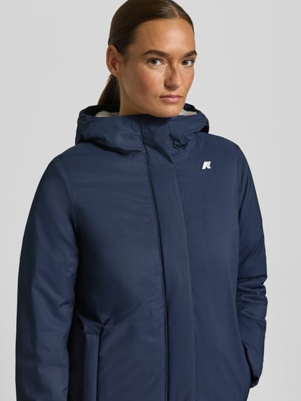 Frauenmodel trägt Regular Fit Mantel mit Logo-Applikation Modell 'SOPHIE' von K-WAY, dunkelblau, glatte Textur, Nahaufnahme Oberkörper. Rückenlänge 73 cm, Schulterbreite 41 cm, Brustweite 46 cm, Ärmellänge 53 cm, Kapuze mit Stehkragen.