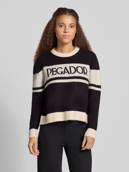 Kobieta modelka nosi sweter z dzianiny z mieszanki materiałów z dodatkiem streczu, krój regular fit, model 'RASSA' od PEGADOR, logo, okrągły dekolt, długość tyłu w rozmiarze S: 55 cm, długość rękawów w rozmiarze S: 54 cm, czarno-kremowe paski, teksturowana powierzchnia, widok całej sylwetki od przodu.