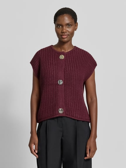 Frauenmodel trägt Relaxed Fit Strickweste mit Woll-Anteil Modell 'Norma' von b.young in Dunkelrot, grobe Strickstruktur, Rundhalsausschnitt, Kappärmel, durchgängige Knopfleiste, Material-Mix, Ganzkörperansicht von vorne.