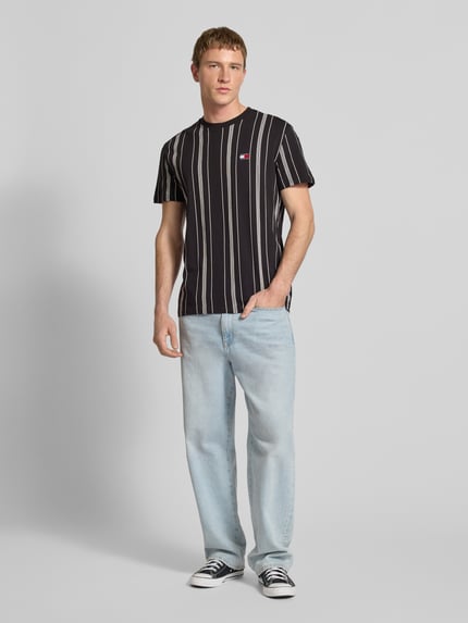 Mannelijk model draagt Regular Fit T-shirt uit puur katoen van Tommy Jeans met all-over motief, regular fit pasvorm, labelstitching applicatie, ronde hals, korte mouw, look: all-over motief, materiaal: puur katoen, kleur zwart met witte strepen, lichtblauwe jeans, Ganzkörperansicht von vorne.