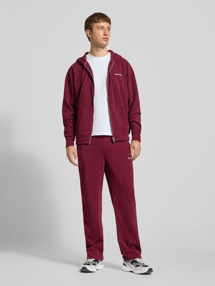 Männermodel trägt Oversized Fit Sweatjacke mit Label-Detail Modell 'Mezzano' von PEGADOR in Dunkelrot, Material-Mix, Ganzkörperansicht von vorne. Rückenlänge bei Größe M: 70 cm, Ärmellänge: 68 cm, elastische Abschlüsse, getrennte Känguru-Tasche.
