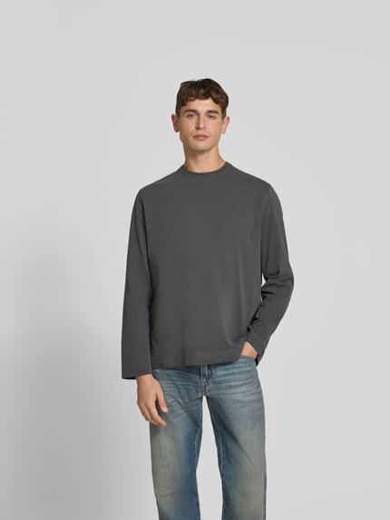 Męski model nosi Relaxed Fit Longsleeve aus Baumwolle von Forét, efekt znoszenia, wyhaftowane logo, okrągły dekolt, długość tyłu 71 cm w rozmiarze M, czarna bawełna, prosty krój, miękka tekstura, Ganzkörperansicht von vorne.
