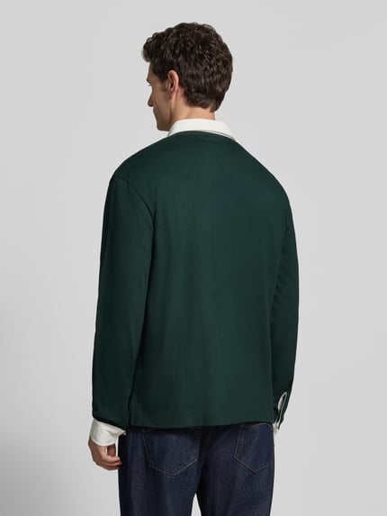 Männermodel trägt Poloshirt mit Logo-Patch von LACOSTE in Dunkelgrün, strukturierte Piqué-Optik, Ganzkörperansicht Rückansicht.