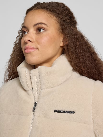 Kobiece model prezentuje Relaxed Fit Jacke in Teddy-Optik Model 'GEORGIA' od PEGADOR, kremowa, imitacja futerka, detal z logo, stójka, zamek błyskawiczny, szerokość w rozmiarze S: 51 cm, długość tyłu w rozmiarze S: 57 cm, szerokość ramion w rozmiarze S: 41 cm, długość rękawów w rozmiarze S: 63 cm, zbliżenie na górną część ciała.