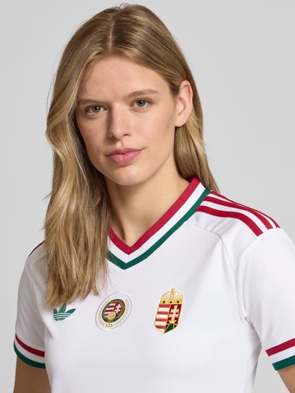 Frauenmodel trägt Ungarn Heimtrikot 2026 von adidas Sportswear in Optik: Unifarben, Regular-Fit, Applikation, Logo-Stitching, Kontraststreifen, V-Ausschnitt, Ärmelform: Kurzarm, Materialverarbeitung: feine Webstruktur, Nahaufnahme Oberkörper.