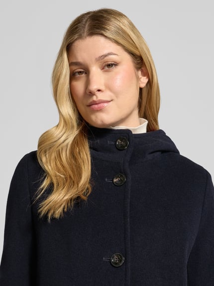 Frauenmodel trägt Wollmantel mit Kapuze von GIL BRET in dunklem Unifarben-Design mit strukturierter Webware, sichtbaren Knöpfen und Kapuze, Nahaufnahme Oberkörper.