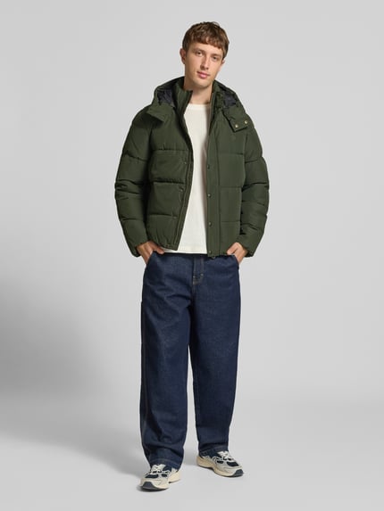 Männermodel trägt Steppjacke mit Kapuze und Reißverschluss von Superdry in Dunkelgrün, Regular Fit, Ganzkörperansicht von vorne. Rückenlänge 71 cm, Schulterbreite 50 cm, Brustweite 56 cm, Ärmellänge 83 cm.
