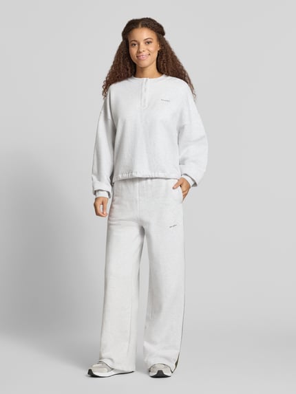 Frauenmodel trägt Boxy Fit Sweatshirt mit Knopfleiste Modell 'Aven' von OH APRIL aus bawełna, okrągły dekolt, wyhaftowane logo, kolor jasnoszary, listewa guzikowa, pudełkowy krój, Ganzkörperansicht von vorne.