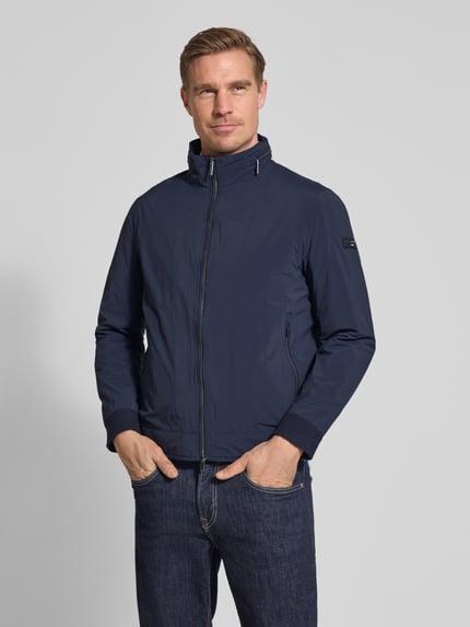 Männermodel trägt Regular Fit Jacke mit Stehkragen von bugatti in Dunkelblau, Zweiwege-Reißverschluss, elastische Ärmelabschlüsse, Eingrifftaschen, Logo-Detail, verstaubare Kapuze, glatte Textur, Ganzkörperansicht von vorne.