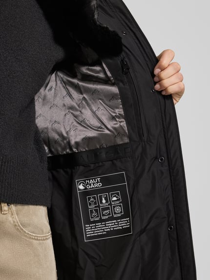 Frauenmodel trägt schwarzen Steppmantel mit Kapuze von NAUTGÅRD 'NEBULA', Innenansicht zeigt Etikett und Futter, Nahaufnahme Oberkörper. Damen Steppmantel Regular Fit mit Kapuze, verdecktem Reißverschluss und Reißverschlusstaschen, Rückenlänge 108 cm, Schulterbreite 42 cm, Brustweite 37 cm, Ärmellänge 67 cm.