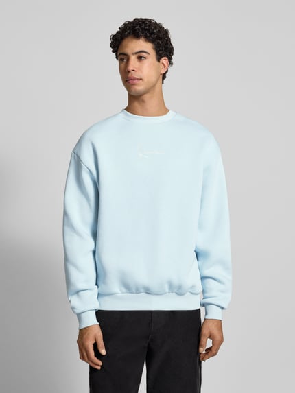 Męski model nosi Sweatshirt mit Label-Stitching od KARL KANI w jasnoniebieskim kolorze, luźny fason, bawełniana mieszanka, zdjęcie całej sylwetki z przodu.