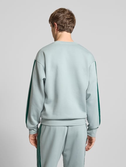 Męski model nosi Sweatshirt mit Rundhalsausschnitt und Logo od adidas Sportswear, bawełniany miks z domieszką wiskozy, krój regular fit, logo, jasnoszary z zielonymi detalami, długość pleców w rozmiarze M: 70 cm, długość rękawa w rozmiarze M: 64 cm, widok całej sylwetki od tyłu