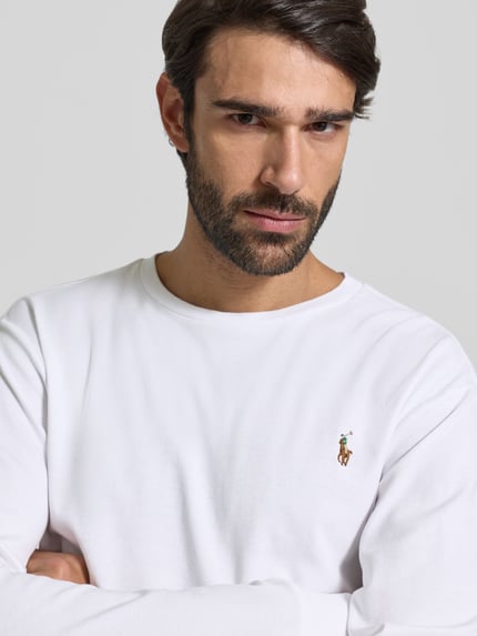 Męski model nosi bluzkę z długim rękawem z prążkowanymi wykończeniami od Polo Ralph Lauren, bawełna, krój regular fit, okrągły dekolt, wyhaftowane logo, długość tyłu 68 cm dla rozmiaru M, jasny kolor, gładka tekstura, zbliżenie na górną część ciała.