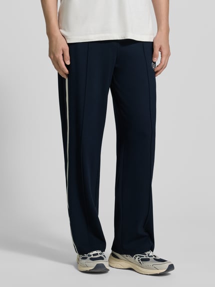 Mężczyzna nosi spodnie typu track pants z mieszanki wiskozy od EA7 Emporio Armani, prosty krój, ozdobne szwy, lampasy, nadruk z logo, wpuszczane kieszenie, pas elastyczny, długość wewnętrzna nogawki w rozmiarze M: 79 cm, zdjęcie całej sylwetki z przodu.