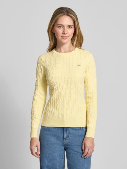 Vrouwenmodel draagt 'Strickpullover mit Zopfmuster und Label-Stitching' van Gant in lichtgeel katoenmix met stretch, slim fit, kabelpatroon, ronde hals, labelstitching, ruglengte 58 cm, mouwlengte 61 cm, volledig zicht van voren.
