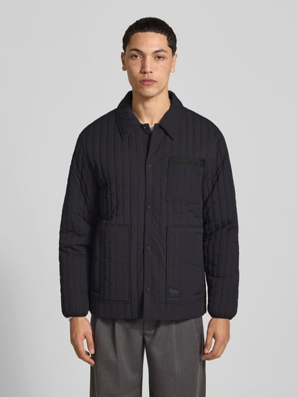 Męski model nosi Steppjacke mit Brusttasche od MAISON KITSUNÉ, czarna, prosty krój, listwa z zatrzaskami, brustasza, wpuszczane kieszenie, detal z logo, elastyczne zakończenia rękawów, długość tyłu w rozmiarze M: 67 cm, szerokość w rozmiarze M: 47 cm, długość rękawów: 62 cm, widok całej sylwetki z przodu.