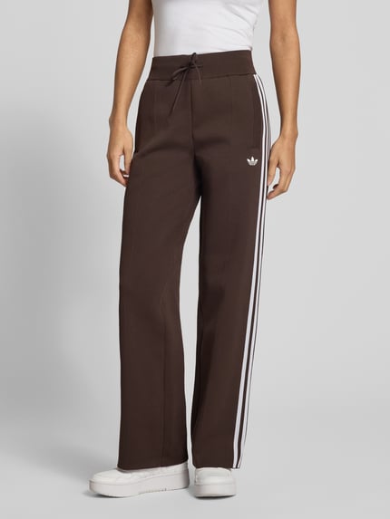 Frauenmodel trägt Regular Fit Sweathose mit Logo-Detail von Adidas Originals in Braun, weiße Streifen, unifarben, seitliche Eingrifftaschen, Tunnelzugbund, Ganzkörperansicht von vorne.