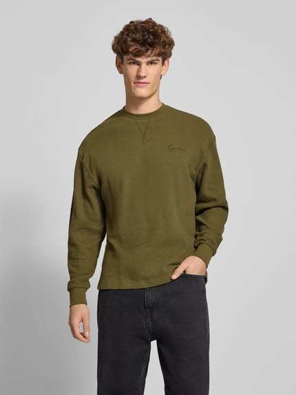 Männermodel trägt Longsleeve mit Label-Stitching von KARL KANI in Olive, Reine Baumwolle, Lockerer-Schnitt, Rundhalsausschnitt, Gerippte-Abschlüsse, Ganzkörperansicht von vorne.